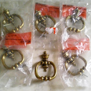 May include: A collection of six brass-toned metal ring pulls for furniture, featuring a circular design with a central mounting post. Five of the pulls are individually sealed in clear plastic packaging with orange labels, while one is loose and unwrapped.