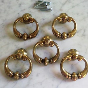 May include: Five antique brass ring pulls with decorative detailing. Each pull has a circular ring and a round backplate. Several screws are visible in the background, suggesting they are ready for installation.