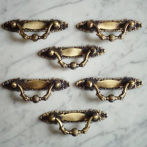 May include: Set of six antique brass cabinet pulls with ornate details and a decorative ring handle. Each pull has a rectangular plate with a slightly raised centre.