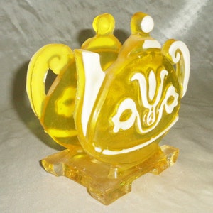 May include: Yellow glass teapot napkin holder with a white design. The holder has a square base.