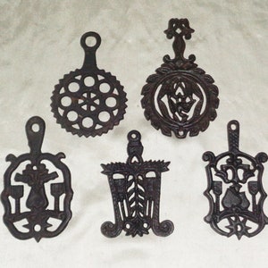 May include: Five cast iron hot pads with intricate designs. The hot pads are black and have a rustic finish. They are all different shapes and sizes, and each has a unique design.