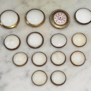 May include: Assortment of 14 round, white glass buttons with gold metal rims. One button has a red and white floral design in the center.