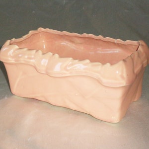 以下が含まれることがあります： テクスチャのあるフリルのある縁が付いた薄いピンクのセラミックプランター。プランターは長方形で、滑らかで光沢のある仕上げになっています。