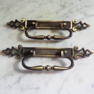 May include: Two antique brass drawer pulls. Each pull features a rectangular centre with a textured wood grain appearance, a curved handle, and decorative end pieces. The pulls are a mix of gold and dark bronze.