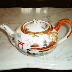 Può includere: Una teiera in porcellana bianca con un design dipinto in rosso e oro che rappresenta un paesaggio giapponese con fiori di ciliegio e una pagoda. La teiera ha un manico marrone e un coperchio con un bordo dorato.