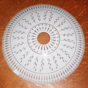May include: A circular white glass light shade featuring a central hole and a decorative pattern of brown and grey painted sprigs, leaves, and radiating diamond shapes arranged in concentric rings across the surface of the glass fixture.