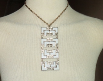 May include: A white and gold geometric pendant necklace. The pendant is made of interlocking squares and rectangles, and it hangs from a gold chain.