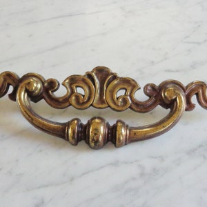 May include: A gold-toned metal cabinet pull with a decorative, ornate design. The pull has a curved shape and a centre knob.