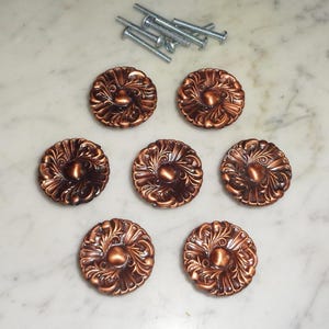 Op de afbeelding: Zeven sierlijke koperen ladegrepen met een bloemmotief. Elke greep heeft een verhoogd midden en een decoratieve rand.