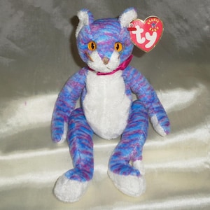 May include: A plush cat toy with blue and purple striped fur, white chest and paws, and orange eyes. It has a pink ribbon around its neck and a heart-shaped tag that says "ty".