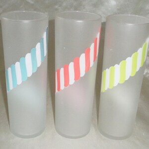 May include: Three frosted glass tumblers with a diagonal stripe design. The tumblers have a white background with blue, pink, and yellow stripes.