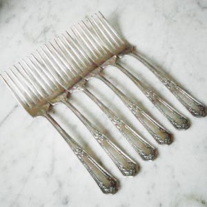 May include: A set of six silver forks with ornate handles. The forks are arranged in a fan shape, with the handles pointing towards the top of the image.