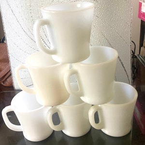 Puede incluir: Seis tazas de cerámica blanca con asas apiladas en forma de pirámide. Las tazas son todas del mismo tamaño y forma, y tienen un acabado liso y brillante.