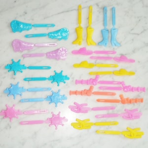 Pode incluir: Um conjunto de 20 grampos de cabelo de plástico coloridos com vários designs de temática western, incluindo botas de cowboy, chapéus, estrelas e instrumentos musicais. Os grampos são em tons de azul, rosa, amarelo e laranja.