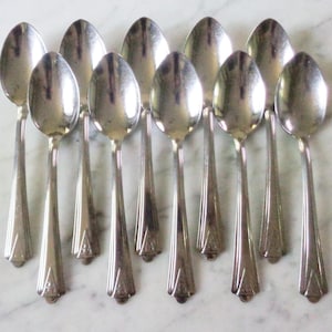 May include: A collection of ten silver spoons arranged in a fan shape. Each spoon has a rounded bowl and a long, tapered handle with a decorative geometric design. The spoons are displayed on a light-colored surface.