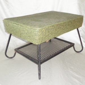 May include: A rectangular ottoman featuring a textured green fabric top with a mottled pattern. The base consists of a black metal frame with four curved legs and a lower shelf made of a black metal mesh grid, providing a stable support structure.