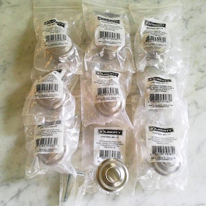 May include: A collection of nine round, silver-toned metal cabinet knobs, each individually packaged in a clear plastic bag. Each bag features a white label with the Liberty brand logo, product code P22782C-SN-CP, and 32 mm diameter.