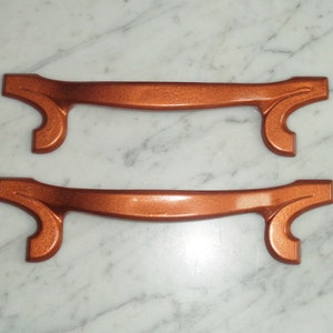 Könnte beinhalten: Zwei kupferfarbene Schranktürgriffe mit einem dekorativen Design. Die Griffe sind aus Metall und haben eine glatte Oberfläche.