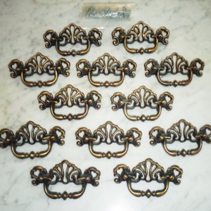 Puede incluir: Un juego de doce tiradores de armario de latón antiguo con un diseño decorativo y ornamentado. Los tiradores tienen una parte superior en forma de abanico y una manija curva.