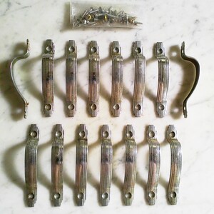 May include: A collection of fourteen silver-toned metal drawer pulls featuring a ribbed, arched design with two screw holes each. The set includes a small plastic bag containing a group of metal mounting screws for installation purposes.