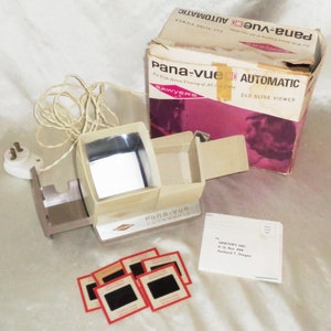 Puede incluir: Un visor de diapositivas 2x2 de plástico beige y marrón con un cable y un enchufe. El visor está etiquetado como "Pana-vue Automatic" y está en su caja original. La caja es rosa y blanca y tiene el texto "Pana-vue Automatic 2x2 Slide Viewer" impreso en ella.