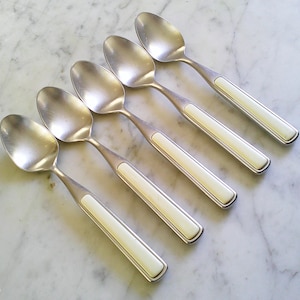 May include: Set of five silver spoons with ivory-colored handles. The spoons are arranged diagonally on a marble surface. The spoons have a classic design, with a rounded bowl and a handle that features a rectangular ivory-colored insert.