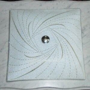 Pode incluir: Uma lumin&aacute;ria de teto quadrada branca com um padr&atilde;o de linhas em espiral e uma pe&ccedil;a central prateada.