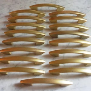 May include: A collection of gold-toned cabinet pulls arranged on a light-colored surface. The pulls have a curved, arched design, and are made of a metallic material. The hardware is likely for furniture or cabinetry.