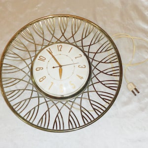 May include: A gold-toned, round, electric clock with a white face and black hands. The clock is surrounded by a decorative, openwork metal frame.