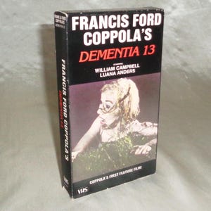 May include: VHS video cassette cover for the film "Dementia 13" directed by Francis Ford Coppola. The cover features a black and white image of a woman with blonde hair in a dark, overgrown area. The text on the cover reads "Francis Ford Coppola's Dementia 13" and "Coppola's First Feature Film!"