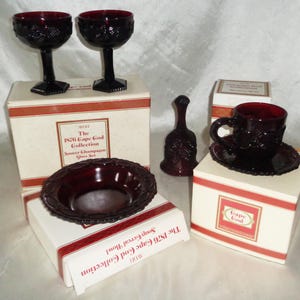 Op de afbeelding: Een verzameling robijnrood glaswerk, waaronder twee champagneglazen, een kleine kom, een bel en een theekopje met schotel. De items worden gepresenteerd op witte dozen met rode accenten en de tekst "The 1976 Cape Cod Collection."