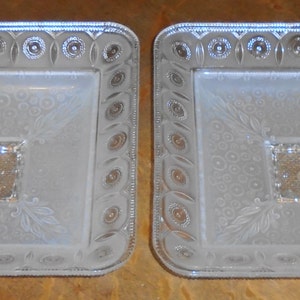 May include: Two clear glass serving dishes with a raised, embossed design. The dishes are square with rounded corners and have a raised center section.