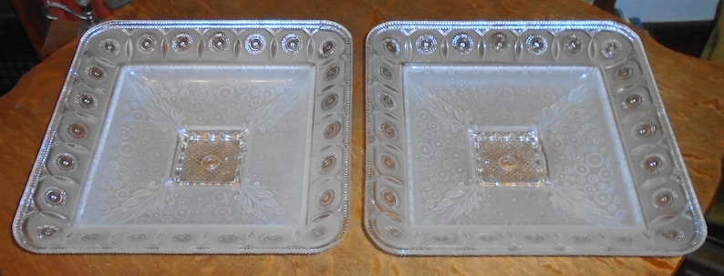 May include: Two clear glass serving dishes with a raised, embossed design. The dishes are square with rounded corners and have a raised center section.