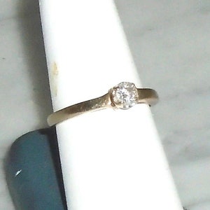 May include: A gold-colored ring with a single, round, clear gemstone. The ring is set on a white display stand. The band is thin and simple, with a slight curve.