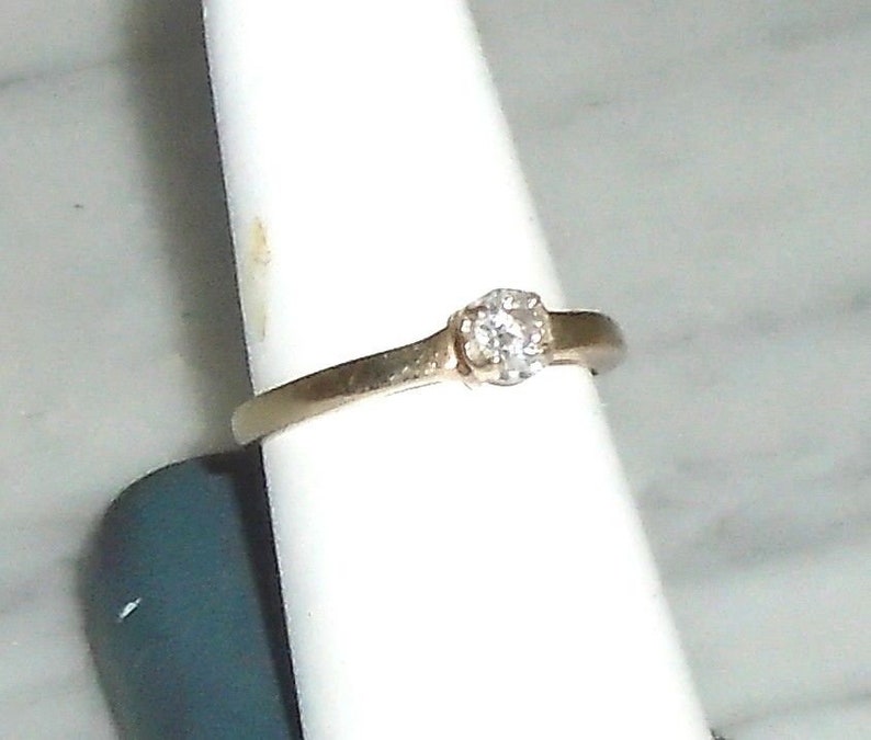 May include: A gold-colored ring with a single, round, clear gemstone. The ring is set on a white display stand. The band is thin and simple, with a slight curve.