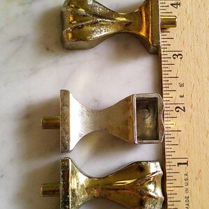 Op de afbeelding: Drie metalen meubelpoten met een goudkleurige afwerking, elk voorzien van een uitlopende voet en een centrale bevestigingspen. Een houten liniaal ligt naast de poten, waaruit blijkt dat elke poot ongeveer 3,8 cm lang is.