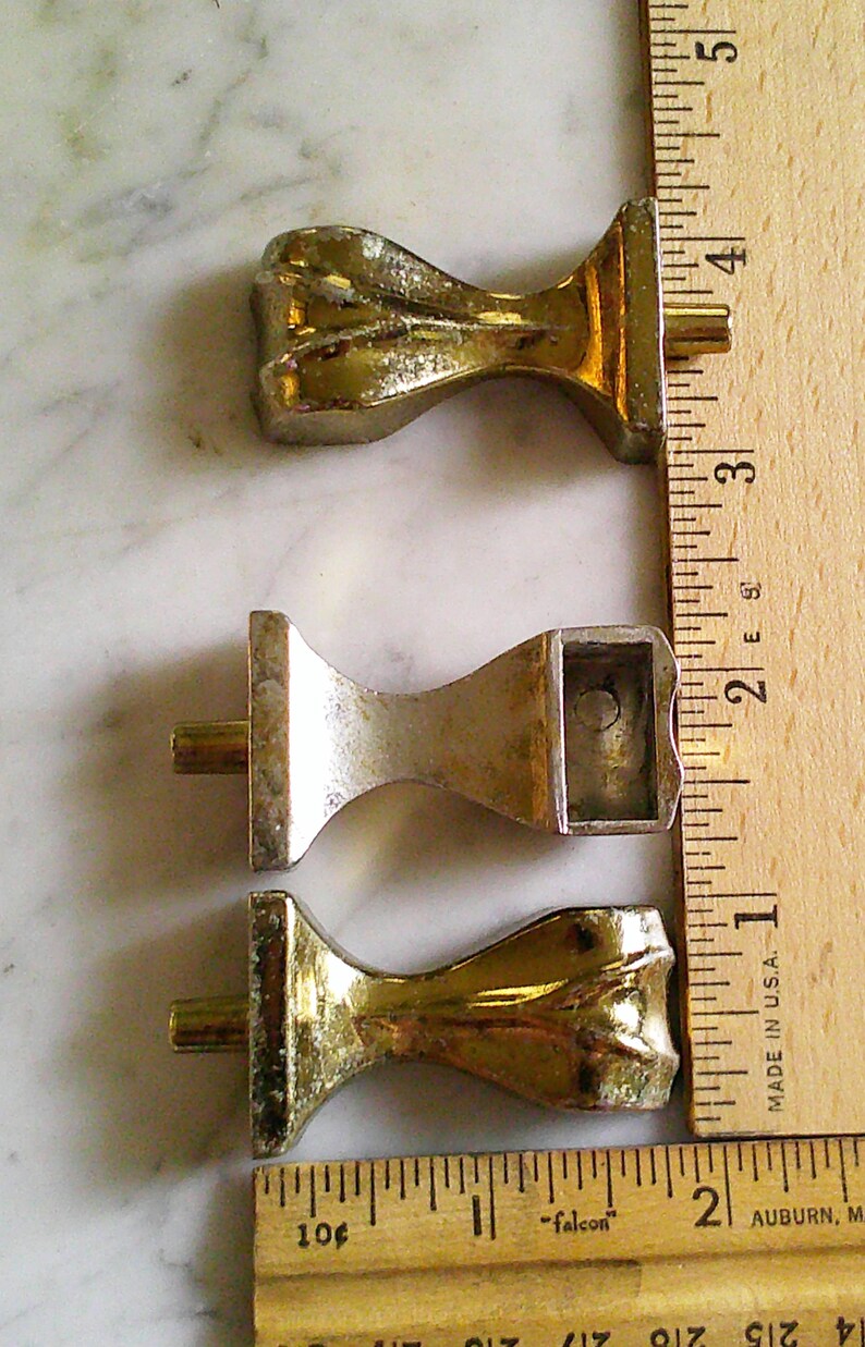 Op de afbeelding: Drie metalen meubelpoten met een goudkleurige afwerking, elk voorzien van een uitlopende voet en een centrale bevestigingspen. Een houten liniaal ligt naast de poten, waaruit blijkt dat elke poot ongeveer 3,8 cm lang is.