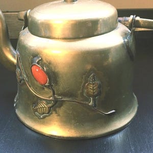 May include: Antique brass teapot with a lid and spout. The teapot features an embossed design with a red oval stone and leaf accents. The teapot is a vintage item, likely used for brewing tea.