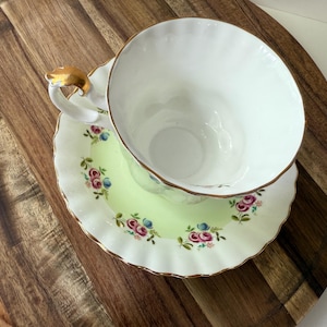 May include: A white teacup with a gold handle sits on a matching saucer. The saucer has a light green center with a floral pattern of pink and blue roses. The rim of the saucer is scalloped and trimmed with gold.