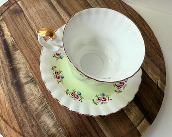 May include: A white teacup with a gold handle sits on a matching saucer. The saucer has a light green center with a floral pattern of pink and blue roses. The rim of the saucer is scalloped and trimmed with gold.