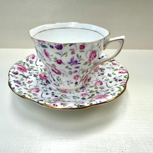 May include: A white teacup and saucer set with a floral pattern in pink, purple, and green. The teacup and saucer have a gold trim. The teacup is on top of the saucer.