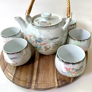 May include: A vintage Japanese tea set featuring a white porcelain teapot with a woven handle and matching cups. The set is decorated with floral and landscape designs in blue, red, and gold, and sits on a wooden serving tray.