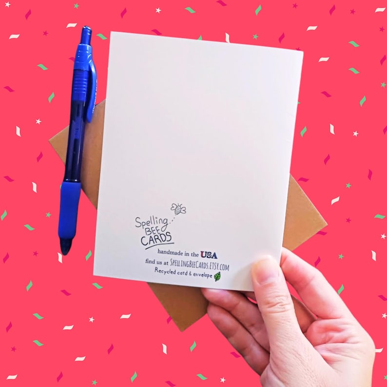 May include: A white card with the text "Spelling Bee Cards" and a bee illustration. The text "handmade in the USA" is below the bee. The text "find us at SPELLINGBEECARDS.ETSY.COM" is below the "handmade in the USA" text. The text "Recycled card & envelope" is below the website address. The card is held in a person's hand.
