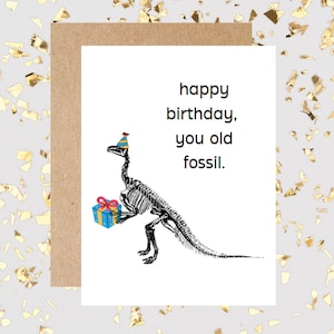 May include: A birthday card with a skeleton dinosaur wearing a party hat and holding a gift. The card reads "happy birthday, you old fossil."