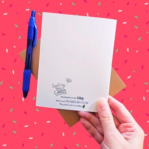 May include: A white card with the text "Spelling Bee Cards" and a bee illustration. The text "handmade in the USA" is below the bee illustration. The text "find us at SPELLINGBEECARDS.ETSY.COM" is below the "handmade in the USA" text. The text "Recycled card & envelope" is below the website address. The card is held in a hand with a brown envelope behind it.