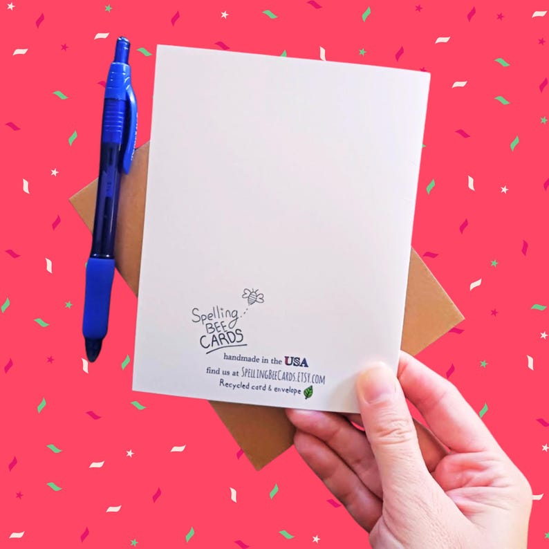 May include: A white card with the text "Spelling Bee Cards" and a bee illustration. The text "handmade in the USA" is below the bee illustration. The text "find us at SPELLINGBEECARDS.ETSY.COM" is below the "handmade in the USA" text. The text "Recycled card & envelope" is below the website address. The card is held in a hand with a brown envelope behind it.