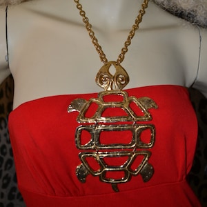May include: A gold chain necklace with a large, ornate turtle pendant. The turtle is made of a textured metal and has a detailed design. The necklace is worn by a person with long, blonde hair and a red strapless dress.