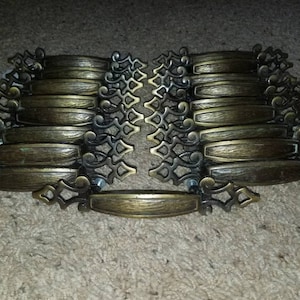 May include: A set of 18 antique brass cabinet drawer pulls with ornate scrollwork and a rectangular plate. The pulls have a brushed finish and are in excellent condition.
