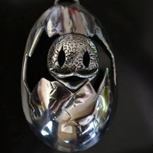 May include: A silver pendant in the shape of a cracked egg with a small, textured creature inside. The creature has two large eyes and a small mouth.