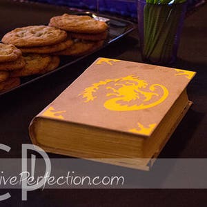 May include: A brown book with a yellow dragon design on the cover. The book is sitting on a black table with a plate of cookies in the background.